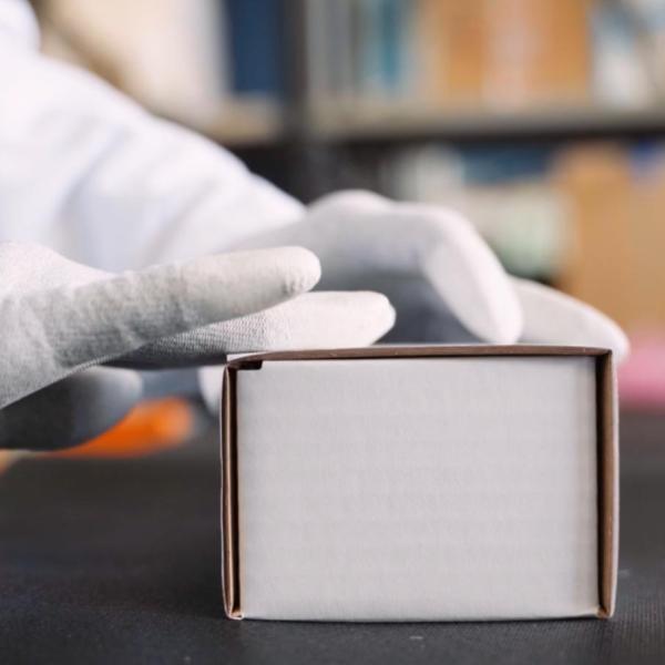 Operatore con guanti protettivi chiude con cura una piccola scatola di cartone su un piano di lavoro.